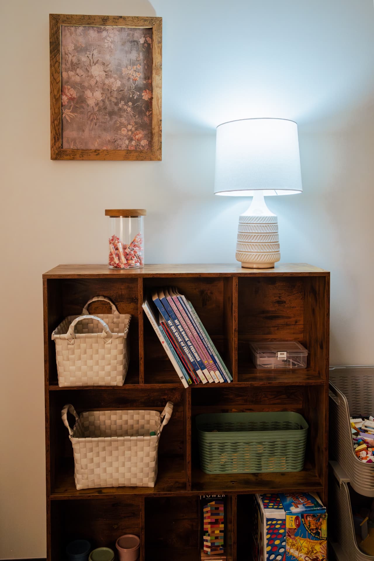 A cozy corner with a bookshelf holding feelings books, baskets, and games beside a warm lamp